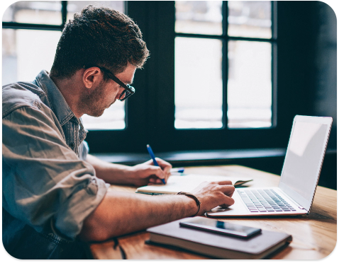 man write in notepad while on laptop