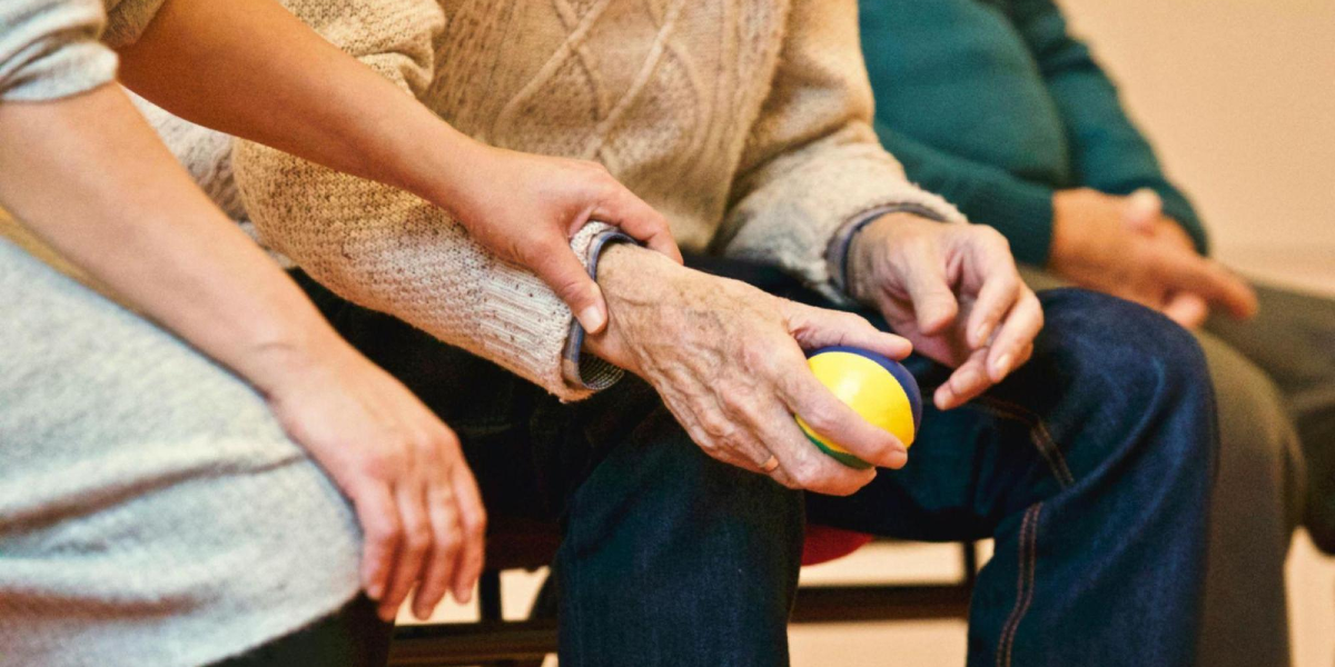 In close-up, a young person places their hand on the wrist of an old person who is holding a ball.