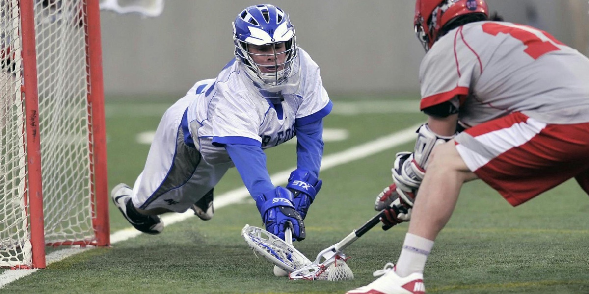 A lacrosse player dives to protect their team's goal as an opposing player attempts to score.
