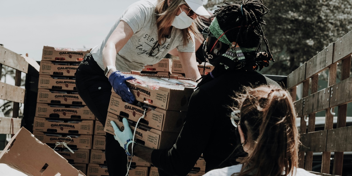 A woman in a white shirt and blue jeans hands a box of food from a truck to another woman.