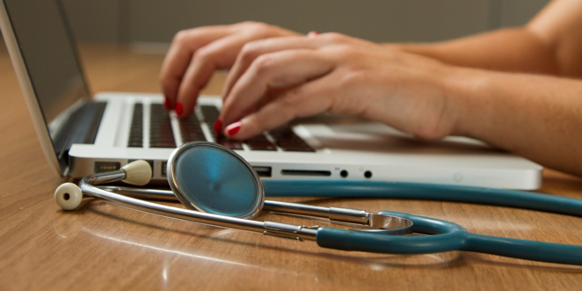 Close-up on a man's hands typing on a laptop with a stethoscope beside it.